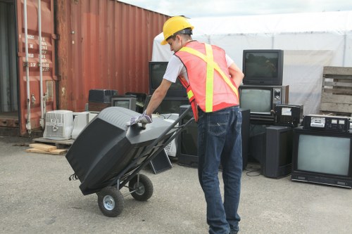 A worker wearing a yellow safety helmet, white shirt, and high-visibility safety vest is pushing a black plastic wheelbarrow filled with rubbish in an outdoor area. The background features a red metal storage container to the left, and several old electronic appliances such as televisions and audio equipment stacked on the right, indicating disposal of electrical waste. The ground is paved, and there is a partially covered structure with a translucent roof behind the worker. The scene suggests a commercial waste removal operation, possibly in Parsons Green, where Commercial Waste Removal Parsons Green offers rubbish collection services. The worker's gloves and the straightforward, functional environment reflect a professional approach to rubbish clearance, with the focus on removing waste from a business or industrial setting.
