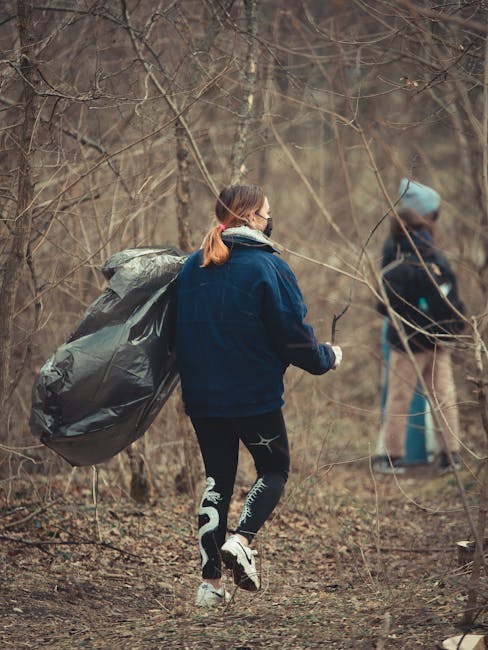 A woman with red hair tied back, dressed in a dark blue jacket, black leggings with white decorative patterns, and sneakers, is walking through a leafless woodland area during daytime while carrying a