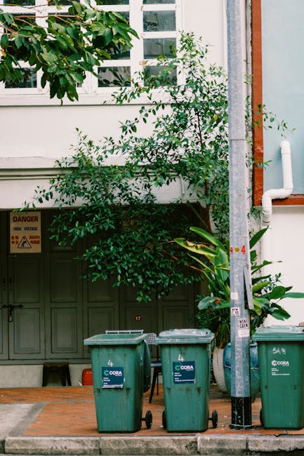 Three green wheelie bins with black lids and white labels are positioned on the sidewalk in front of a building with a gray garage door. Behind the bins, a leafy shrub with dark green foliage partiall